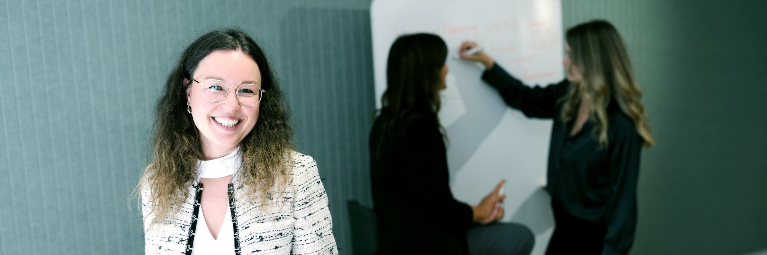 Auf diesem Bild ist im Vordergrund Pia-Maria Aichinger zusehen, die in die Kamera lächelt und im Hintergrund schreiben zwei Kolleginnen an einem Whiteboard.