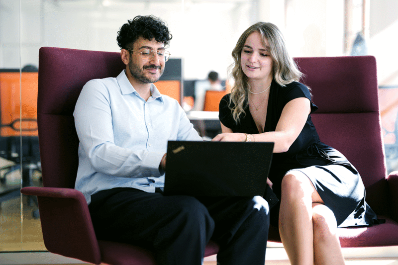 Two consultants are sitting on red chairs with a laptop in front of them. the female on the right is showing the male on the left something on the laptop.