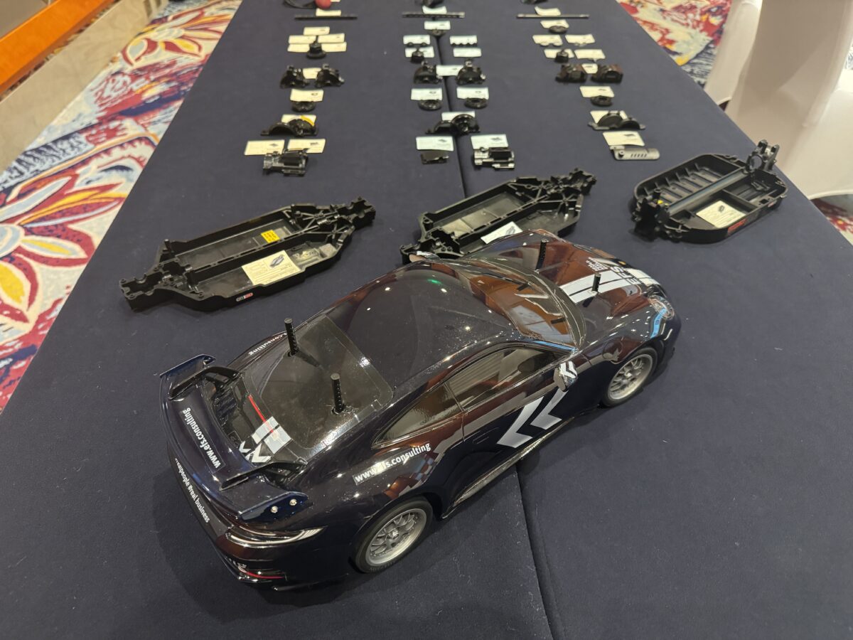This photo shows a black model vehicle with gray arrows on the side, standing on a table with many other labeled components.