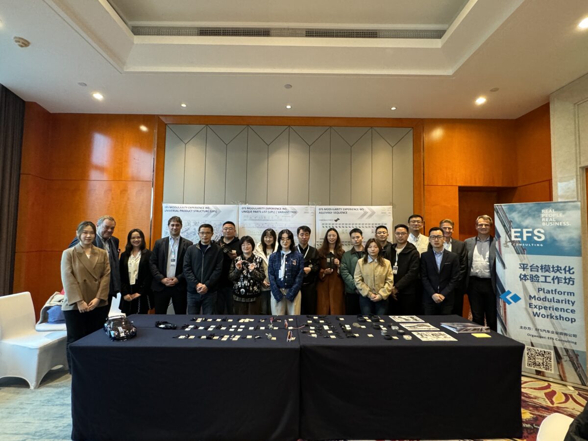 This picture shows a group of people standing behind a table covered with labeled components, all smiling. To the right of the group is a roll-up banner with the EFS Consulting logo on a light blue background. Three white posters can be seen on the wall behind them.