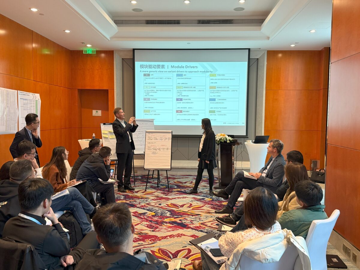 This photo shows a circle of people sitting down. In the middle stands a man in a suit next to a woman dressed in black, who are looking at a PowerPoint slide projection above them and explaining something.