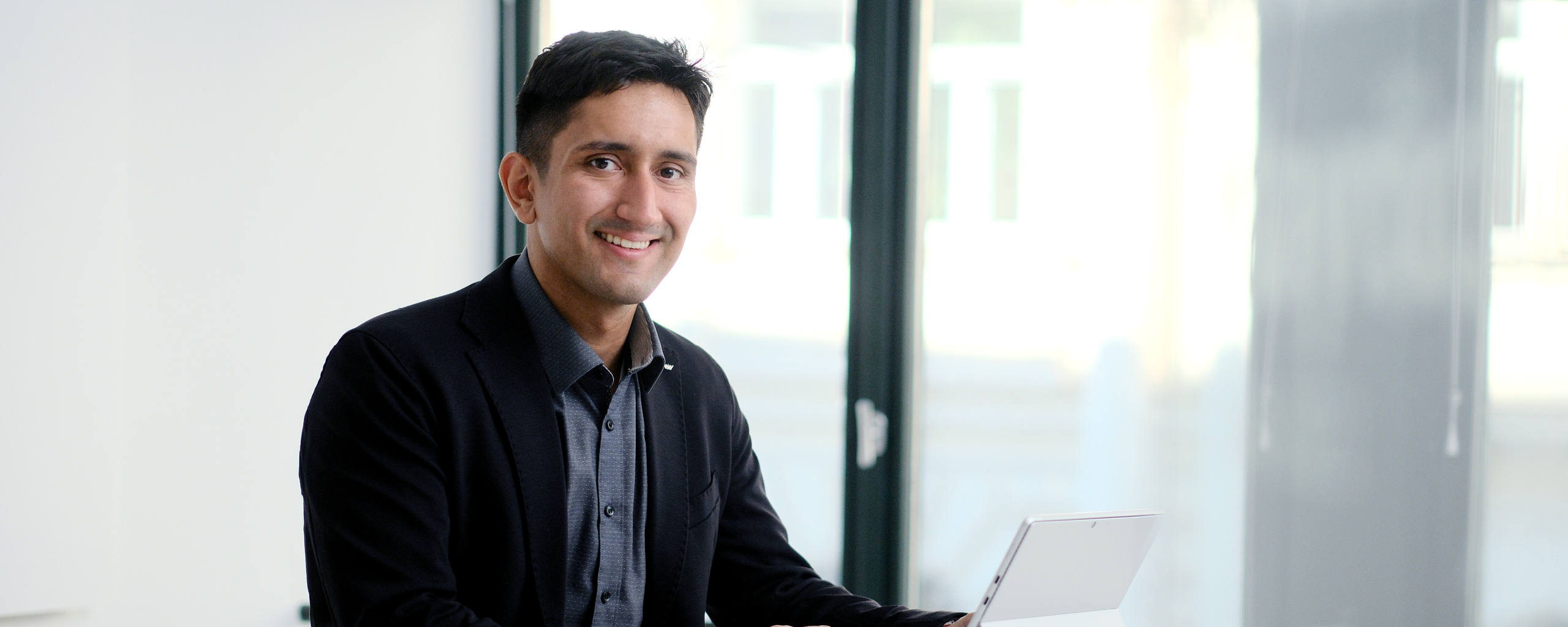 A consultant in a suit sits in front of a laptop smiling in a bright, modern office.
