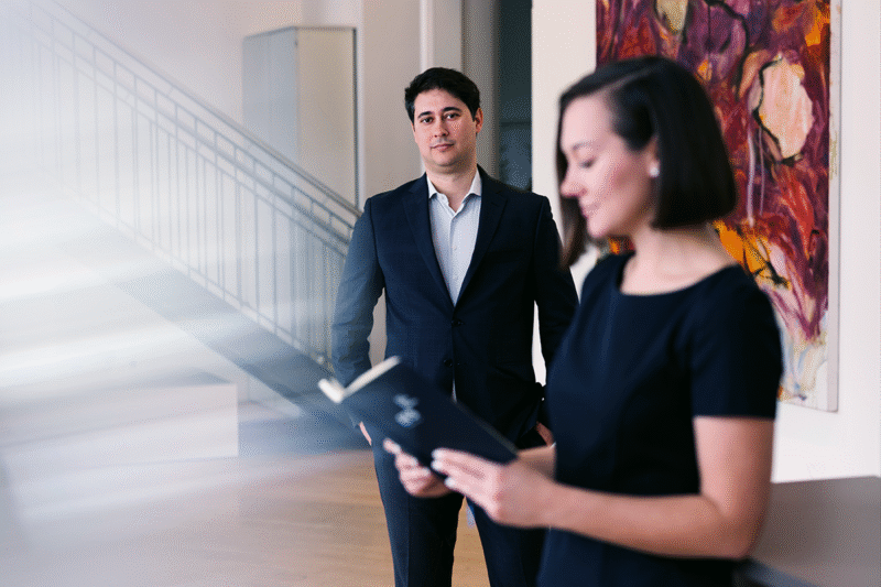 Beraterin mit Buch im hellen Büro im Fokus, Berater im Hintergrund; rotes Wandbild und Treppe als Setting.