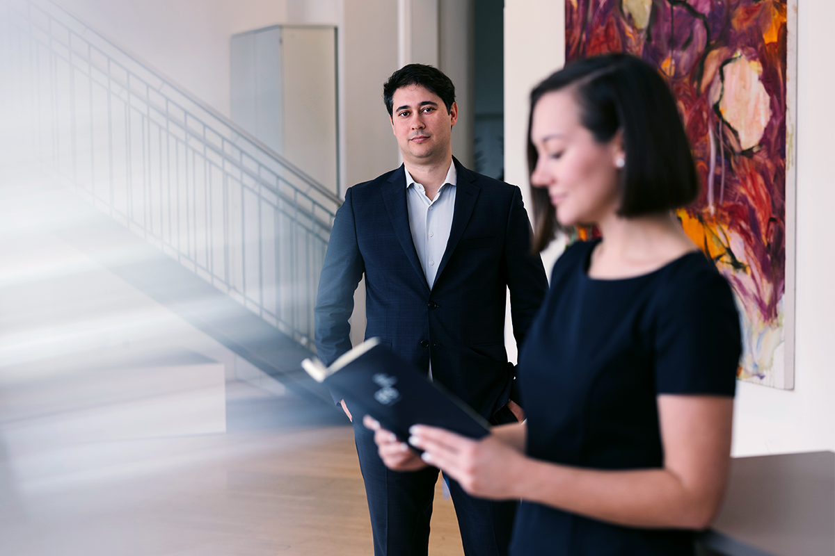 Beraterin mit Buch im hellen Büro im Fokus, Berater im Hintergrund; rotes Wandbild und Treppe als Setting.