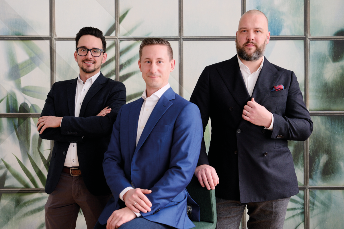 This picture shows four men standing in a row in front of a green wall, smiling at the camera.