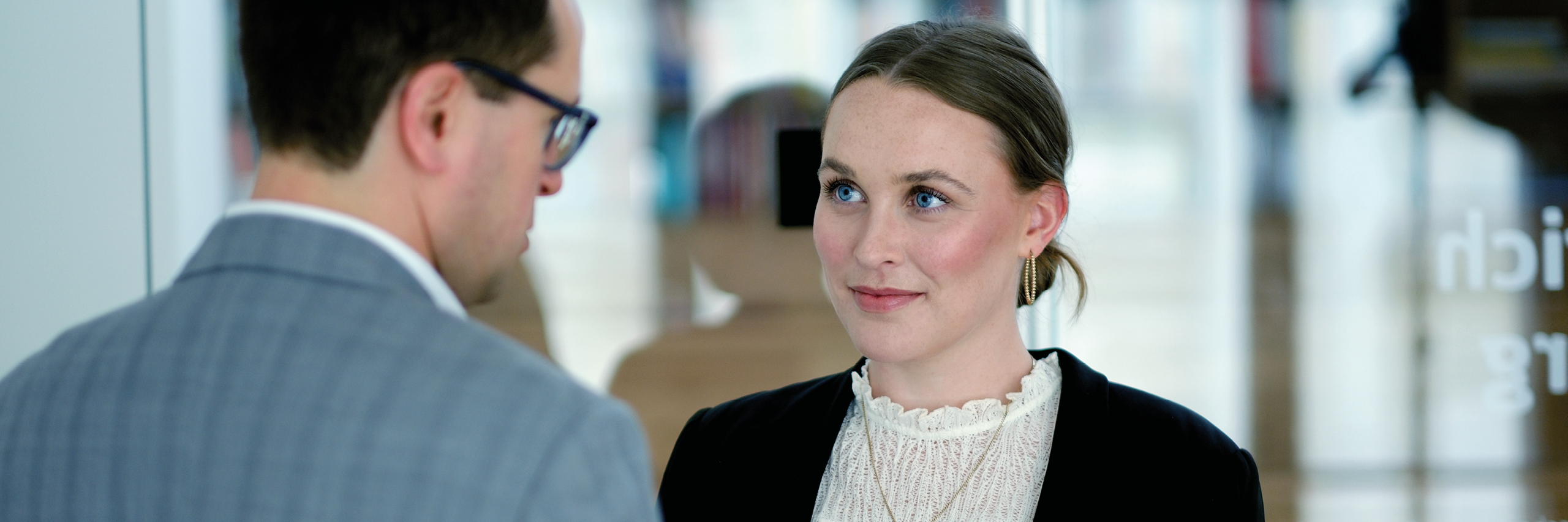 Two people engaged in a conversation. A woman with light skin, blue eyes, and hair tied back is in focus. She is wearing a black jacket over a white lace blouse and gold hoop earrings as she looks at the man opposite her. The man, seen mostly from the back and side, is wearing glasses and a light gray checkered suit. The background is softly blurred, suggesting an indoor office or professional setting with glass walls and some indistinct shapes behind them.