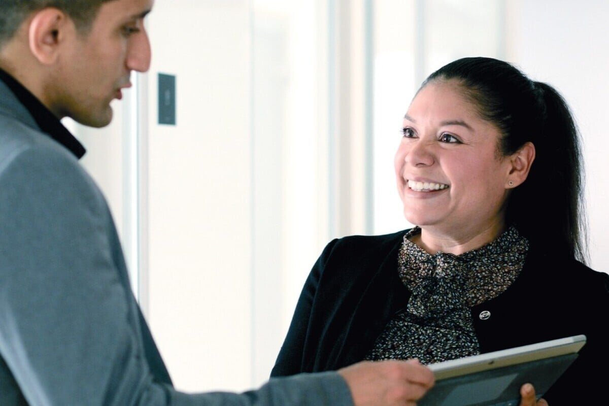 Freundliche Mitarbeiterin hält ein Tablet und spricht mit einem Kollegen in einem hellen Büroflur.