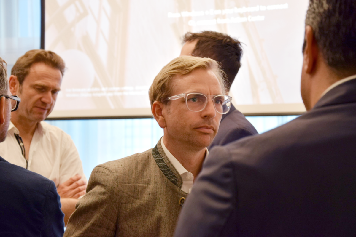 Mehrere Personen stehen in Gruppen beieinander und unterhalten sich bei einer Veranstaltung; im Vordergrund ist Felix Meizer mit Brille im Gespräch, im Hintergrund eine Projektionsfläche.