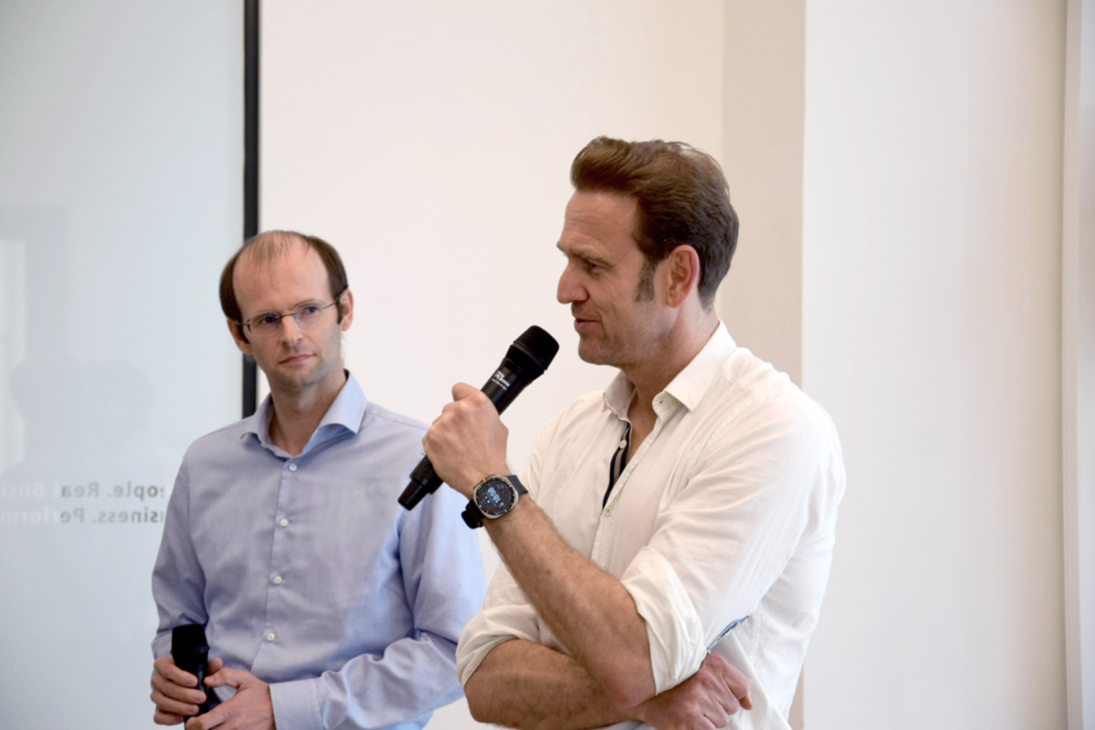 Jochen Schreiber und Julian Kawohl stehen vor einer Projektionsfläche; eine Person spricht in ein Handmikrofon, während die andere daneben zuhört und ebenfalls ein Mikrofon hält.